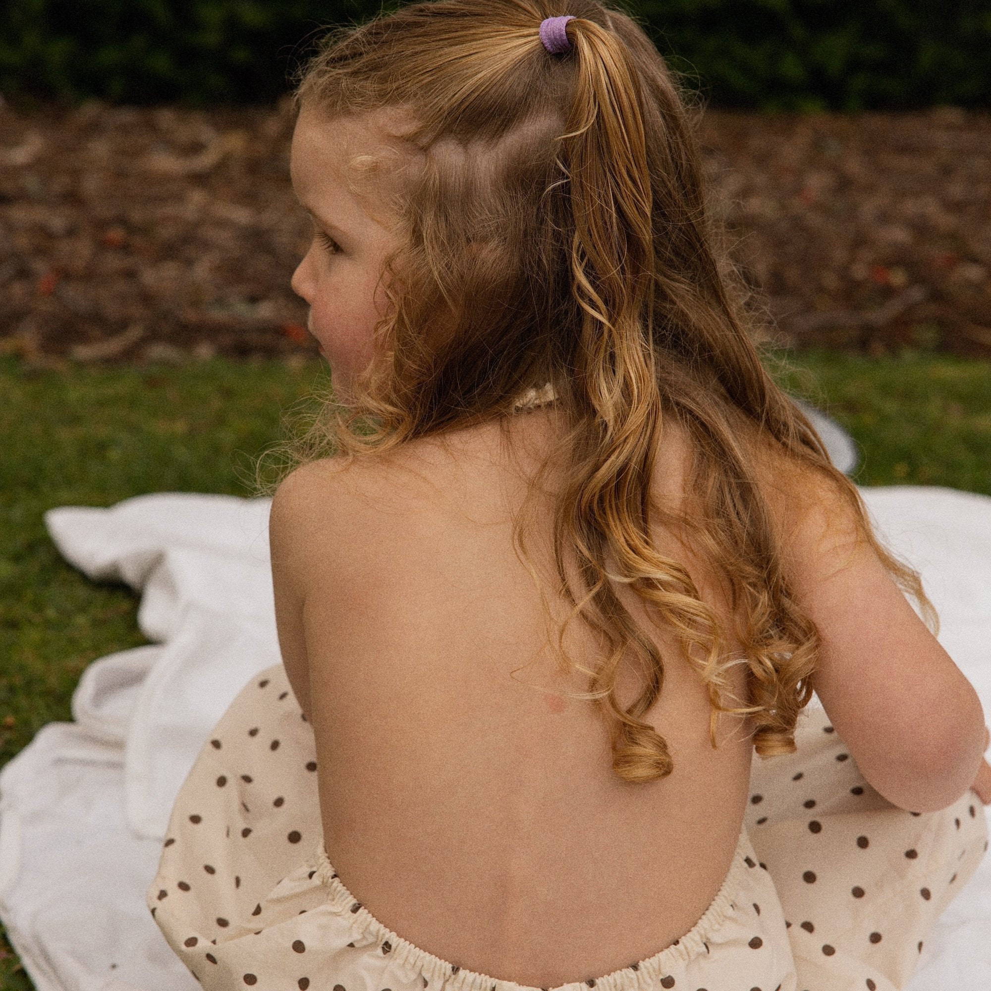 Child sitting on a picnic blanket in the Daisy Dot Jumpsuit, showing the scoop back and gathered waist
