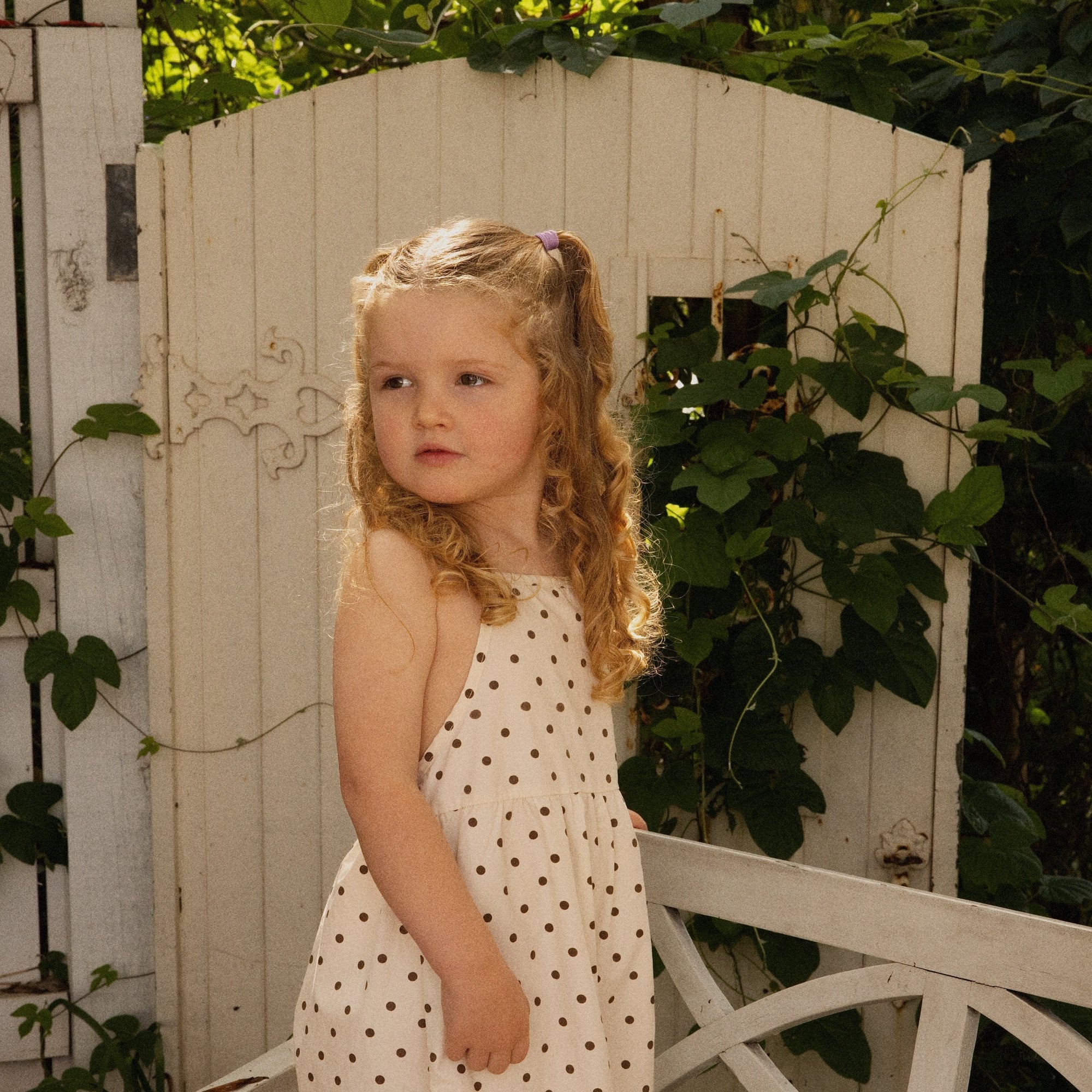 Child at a garden gate in the Daisy Dot Jumpsuit in cream with brown spots