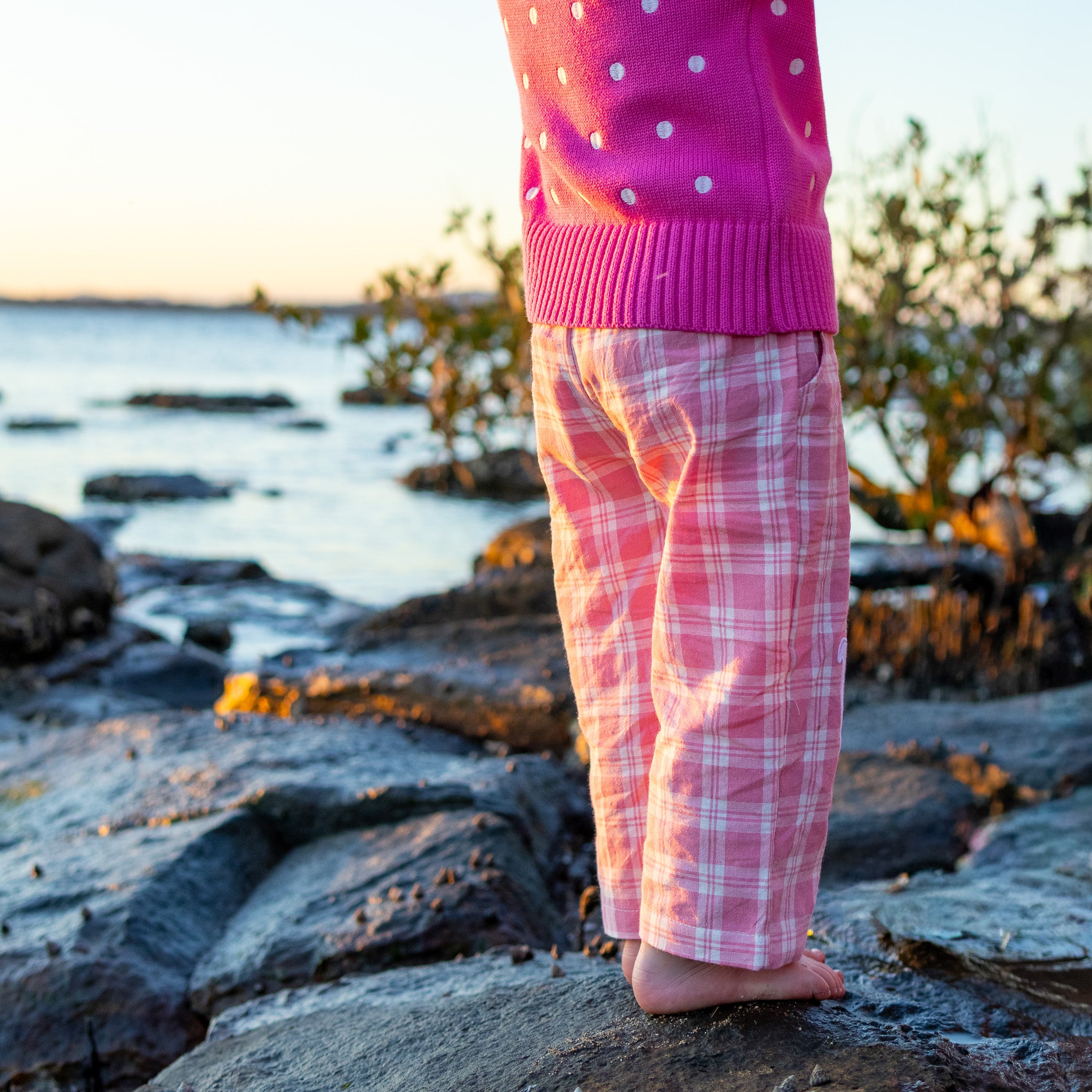 Little Dolphin Club Pants in pink check worn with a pink spot cardi, back view on the rocks at sunset