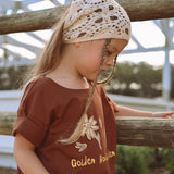 Child leaning on a wooden fence at golden hour wearing the Golden Hour Garden Tee in brown, showing the hand drawn flower and bee print