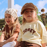 Two children sitting in the sun, one wearing the Sweet as Honey Tee in pale butter yellow with the HONEY drip print across the front