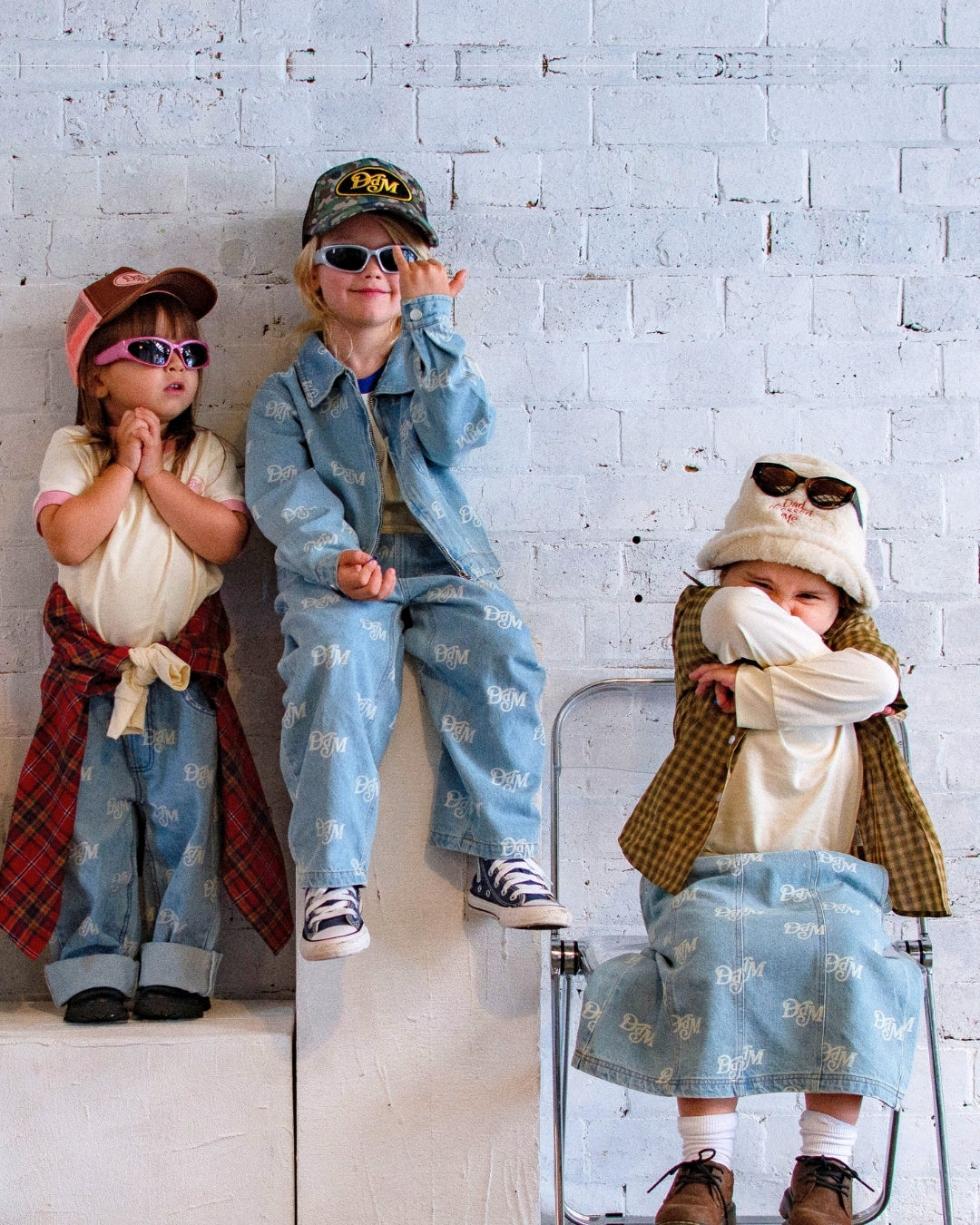 Three children in stylish outfits against a white brick wall.