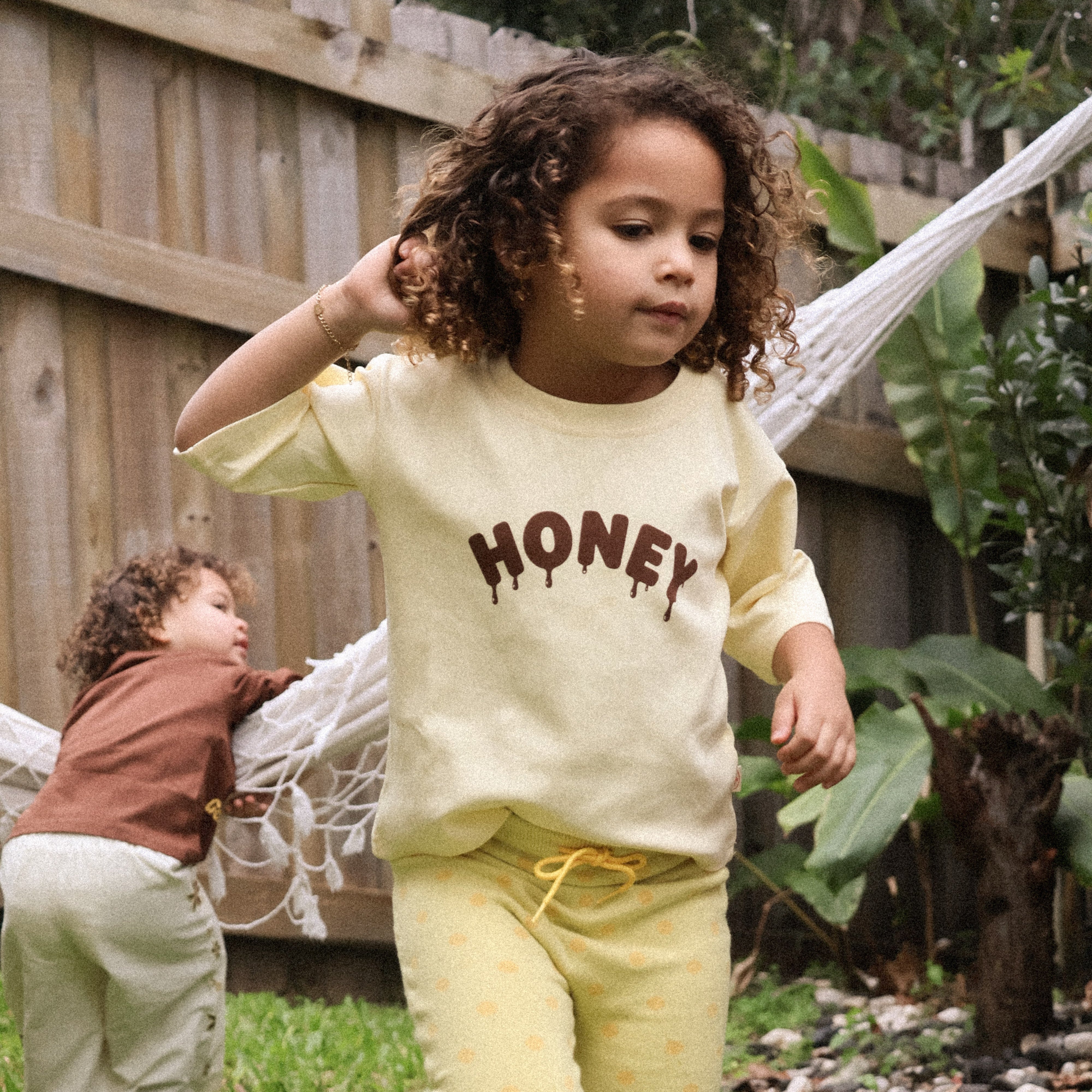 Child running through a garden in the Sweet as Honey Tee and Buttercup Flares, hammock behind
