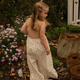 Child standing among tree stumps and pink daisies at golden hour, wearing the Daisy Dot Jumpsuit from the Golden Hour Garden collection