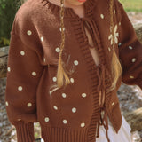 Child wearing the Honey Blossom Cardi in chocolate brown knit with cream polka dots, tie-up front and flower detail on the sleeve, paired with a white skirt